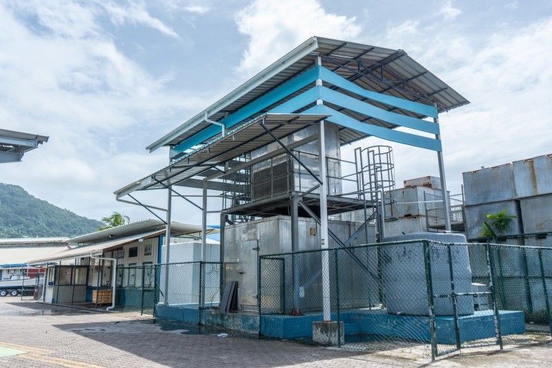 A blue and white industrial structure with a metal roof, likely an ice plant, behind a chain-link fence.