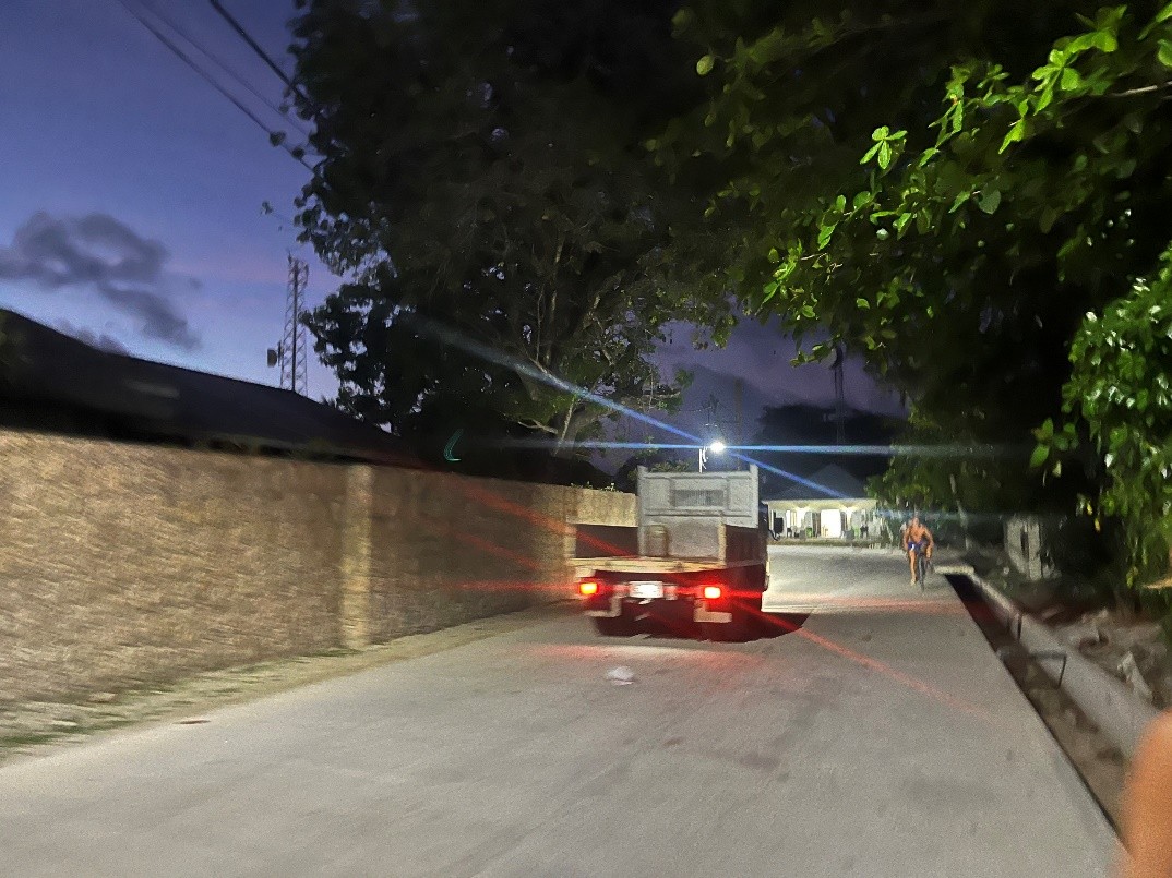 A truck driving down a road at dusk with a stone wall on one side and trees on the other.