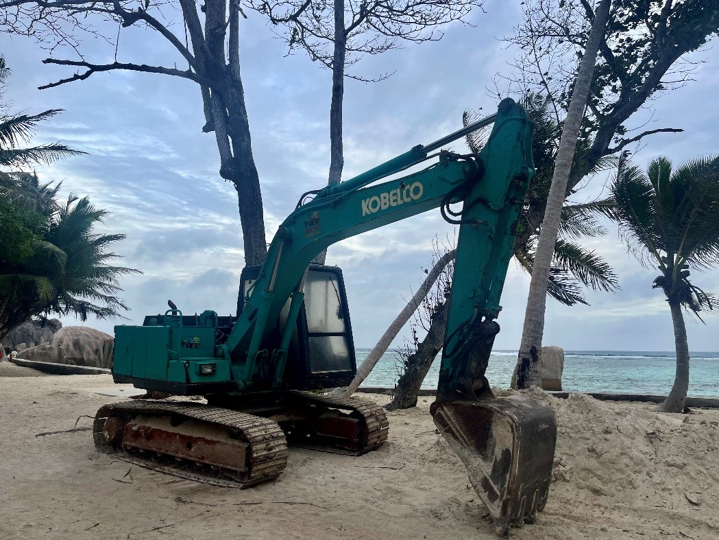An excavator parked on a sandy beach with palm trees and the ocean behind it.